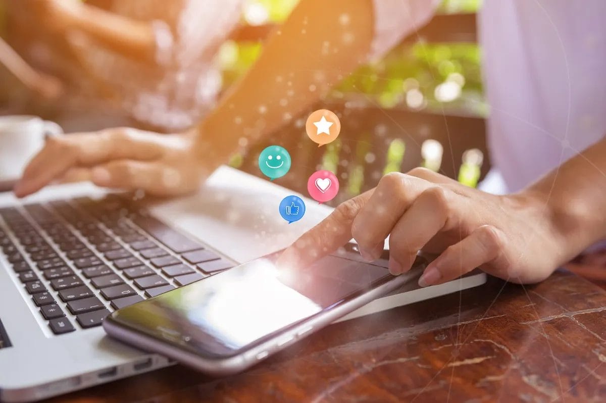 Hands using laptop and phone with social media icons.