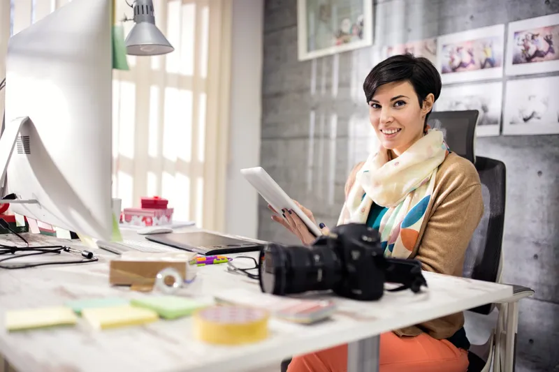 Woman smiling at desk with tablet and camera.
